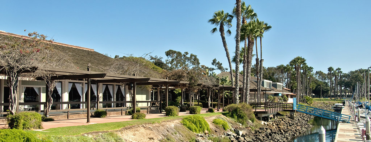 Waterfront restaurant terrace beside a marina dock with boats and palm trees, likely a Southern California harbor resort.
