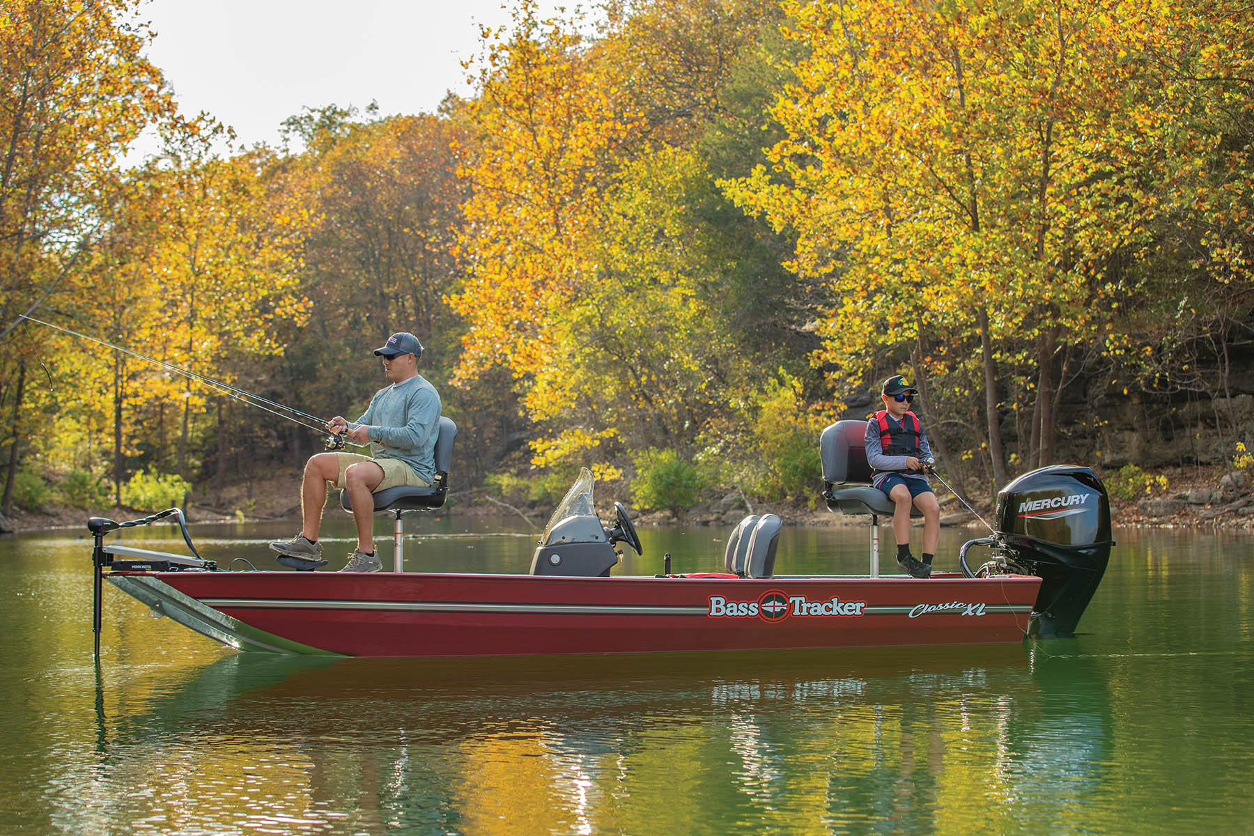 Red Bass Tracker Classic XL fishing boat with a Mercury outboard motor, carrying two anglers fishing on a lake lined with autumn foliage.