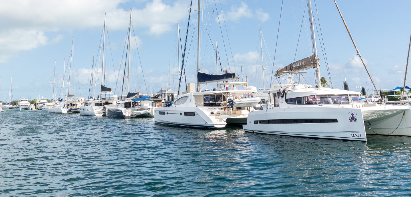 Catamarans and sailboats moored along the docks of a marina basin under a partly cloudy sky.