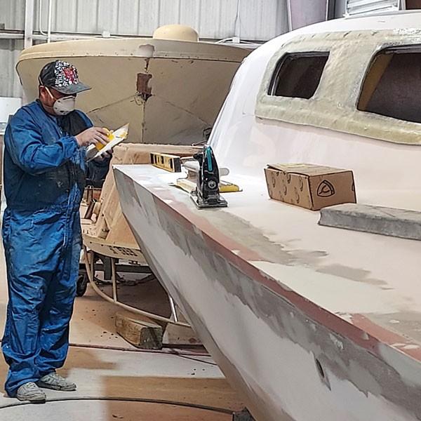 Technician sanding and prepping a white fiberglass boat hull with a power sander inside a boat shop.