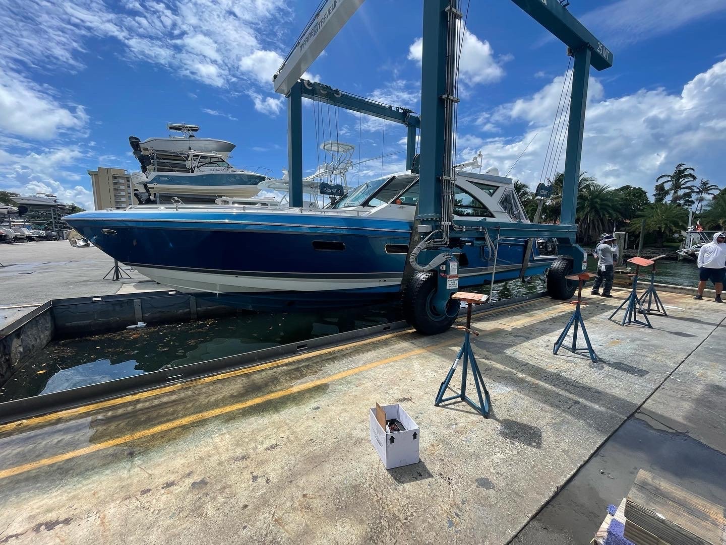 Blue motor yacht being hoisted on a 25-ton travel lift at a boatyard with support stands on the concrete pad.