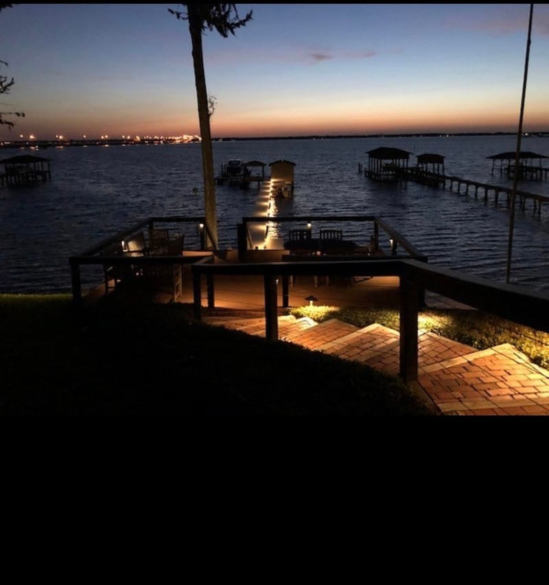 Waterfront deck and lit brick steps leading to a dock with covered boat slips on the water at dusk.