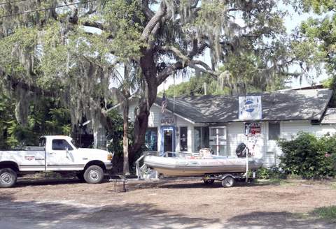 Inflatable boat on a trailer in front of a small marine shop under a moss-draped oak tree.
