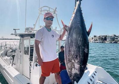 Angler holding up a large tuna on a sportfishing boat in a harbor with waterfront homes in the background.
