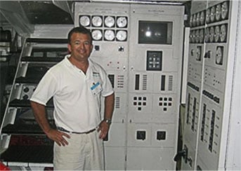 Crew member standing beside a vessel's electrical switchboard and control panels in an engine room.
