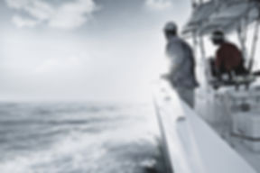 Two crew members standing on the deck rail of a boat while it moves across choppy open water.