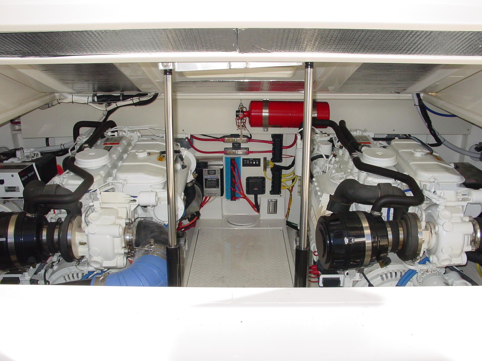 Twin diesel marine engines with a red fire suppression cylinder and wiring panel in a yacht engine compartment.