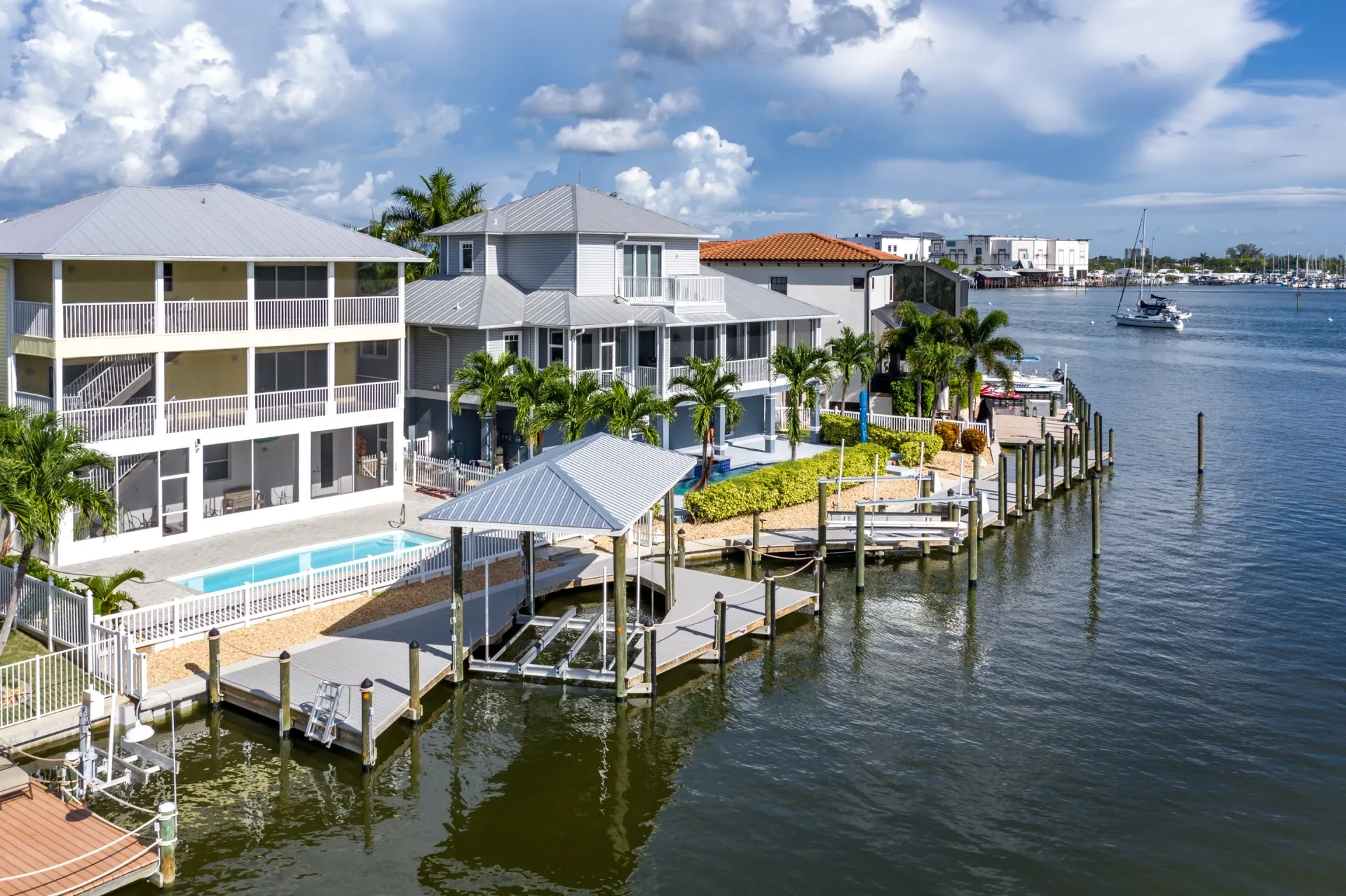 Waterfront homes with private docks, a covered boat lift, and pools along a canal with a sailboat in the background.