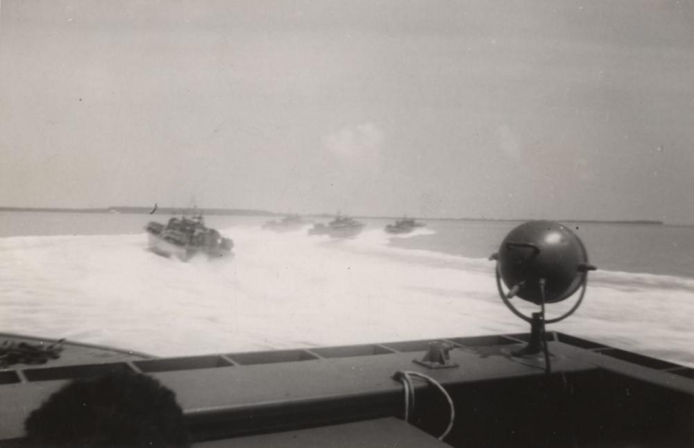 Several landing craft speeding in formation across open water, photographed from the bow of a boat with a spherical searchlight mounted on the rail.