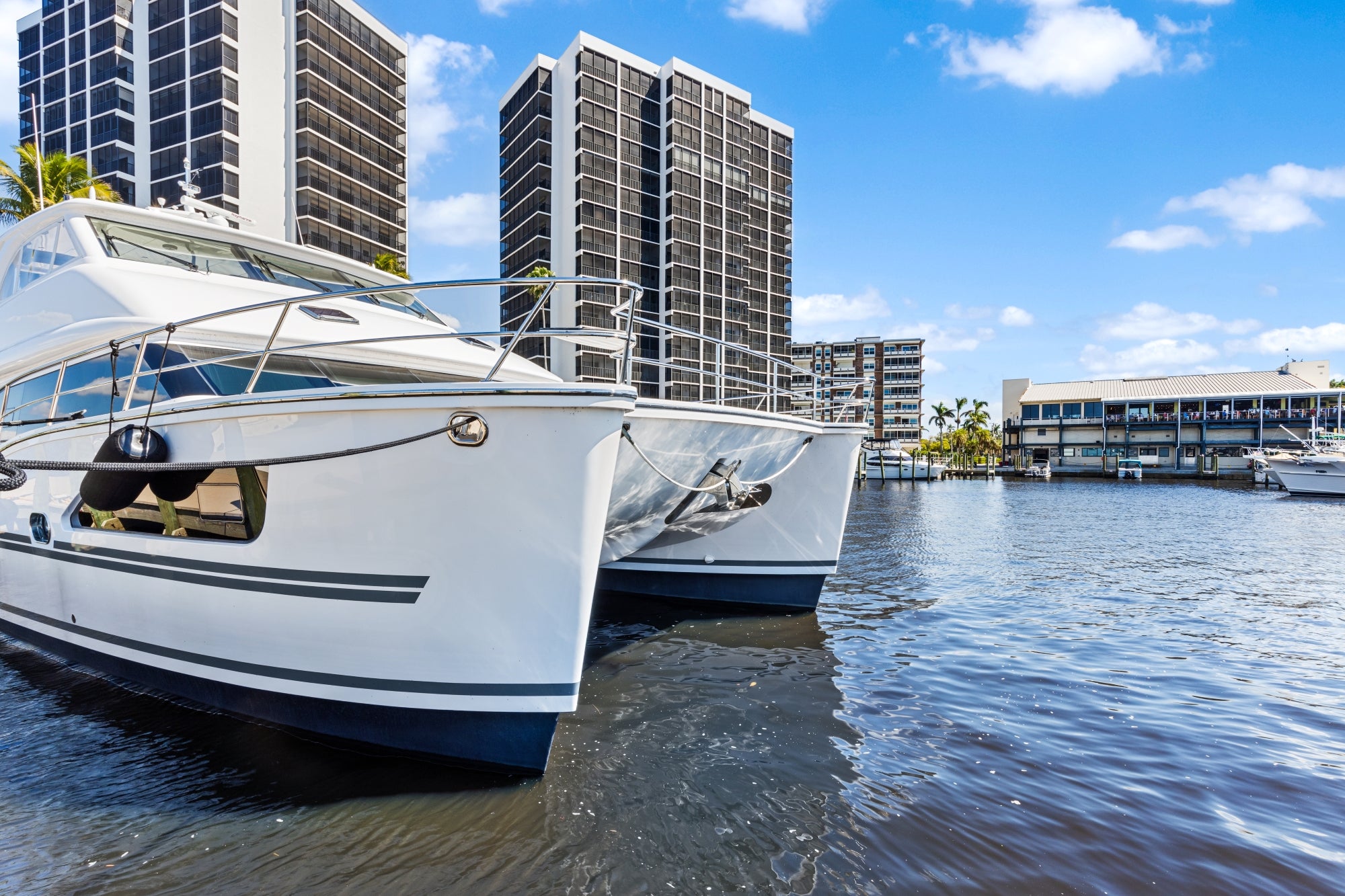 Two white motor yachts moored along a canal with high-rise condominiums and a waterfront restaurant with docks behind them.