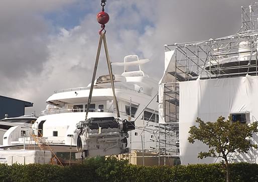 Large white motor yacht with crane-lifted equipment and scaffolding at a boatyard.