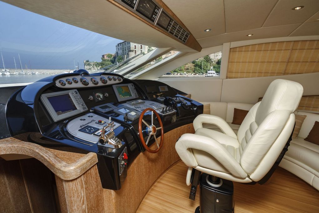 Helm station with wheel, engine controls and navigation displays on a motor yacht, with a marina visible through the windows.