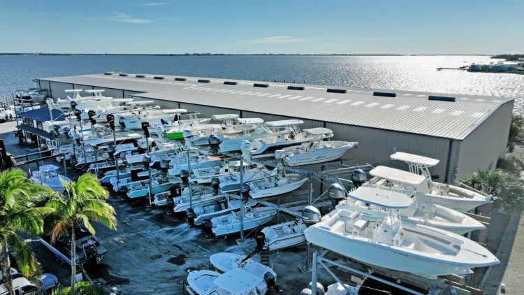 Aerial view of sport fishing boats in outdoor storage beside a metal storage building on the waterfront, with palm trees and a bay in the background.