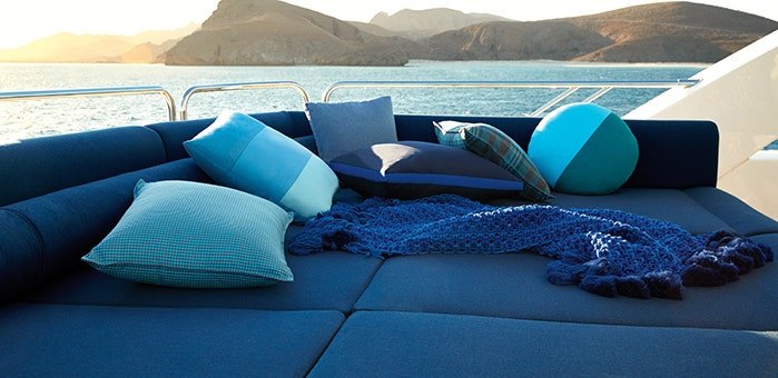Blue marine upholstery, cushions and knit throw on a boat's aft lounge with coastal mountains and calm water beyond.