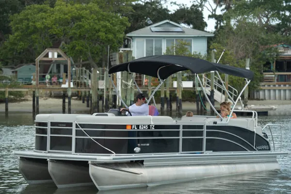 Pontoon boat with a black bimini top carrying passengers on calm water near waterfront homes, registered in Florida (FL 8245 TZ).