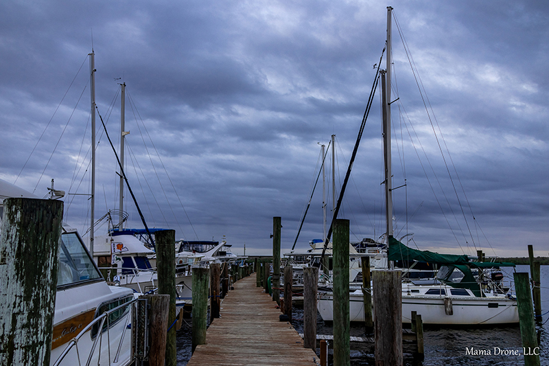 Wooden dock walkway with moored sailboats and motorboats under a cloudy sky, with a Mama Drone, LLC watermark.