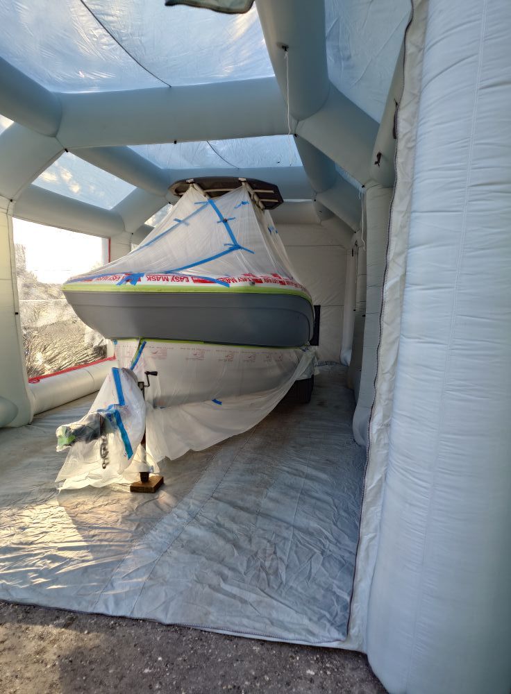 A grey and white motorboat masked off with painter's tape and plastic sheeting inside an inflatable spray booth tent, prepared for coating work.