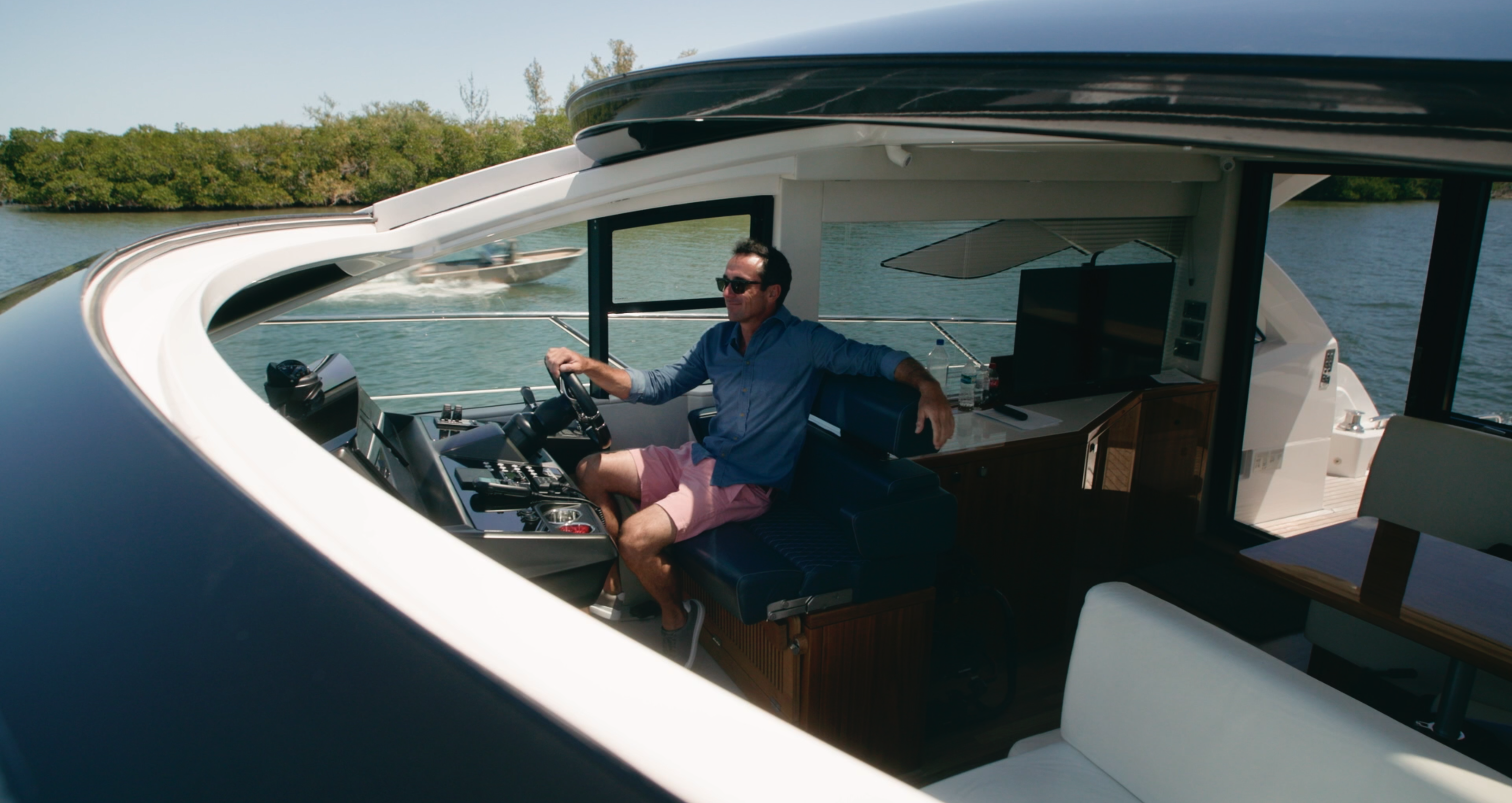 Man at the helm of a yacht with a navy and white hull cruising past mangroves while another boat passes nearby.