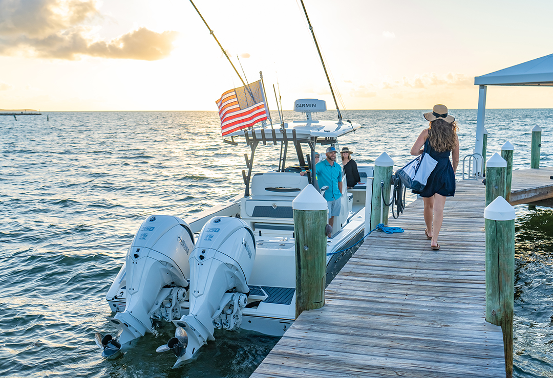 Center console boat with twin outboard motors docked at a wooden pier at sunset, with passengers boarding.