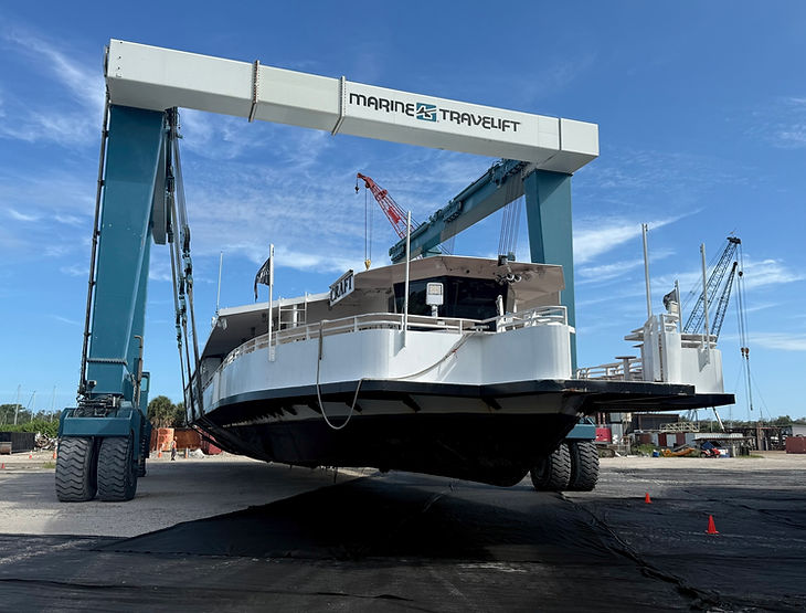 White workboat being lifted by a Marine Travelift gantry at a boatyard with cranes and yard equipment in the background.