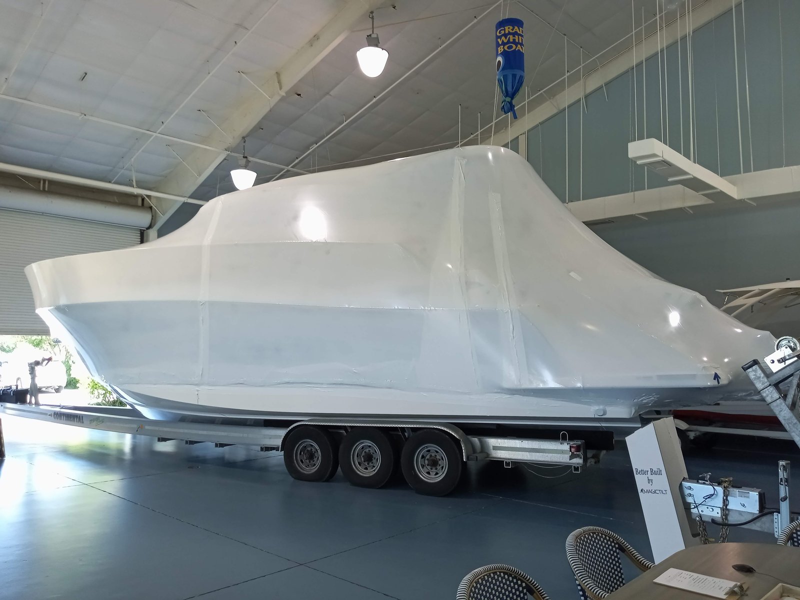 Large white fiberglass boat hull wrapped in protective film on a Continental trailer at a Grady-White dealership showroom, with a MagicTilt sign nearby.