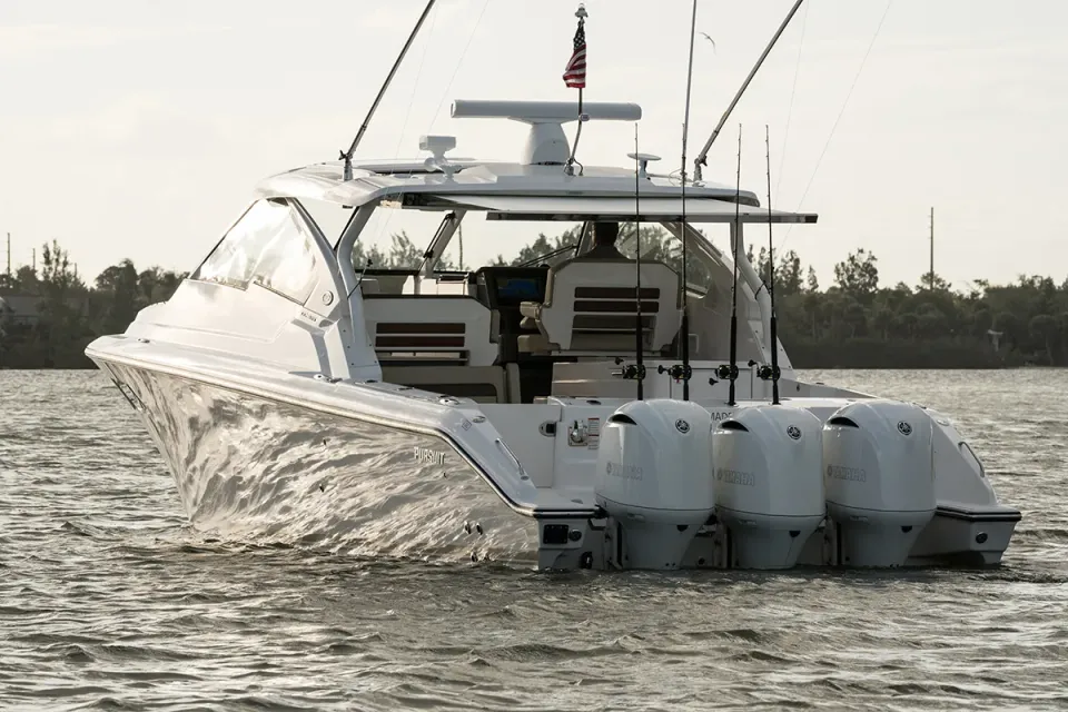 White sport fishing boat with three outboard engines and rod holders running on open water, with a tree-lined shoreline behind it.