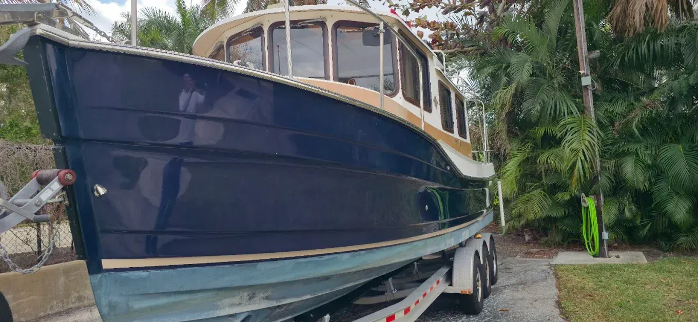 Navy-hulled trawler-style motor yacht with cream and tan trim resting on a trailer in a palm-lined yard.