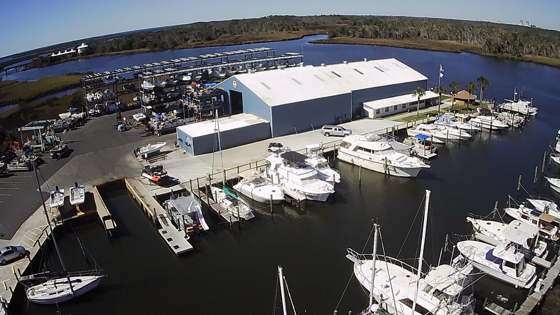 Aerial view of a boatyard with a large storage and service hall, dry-stack boat racks, and yachts moored at docks along a tidal creek.