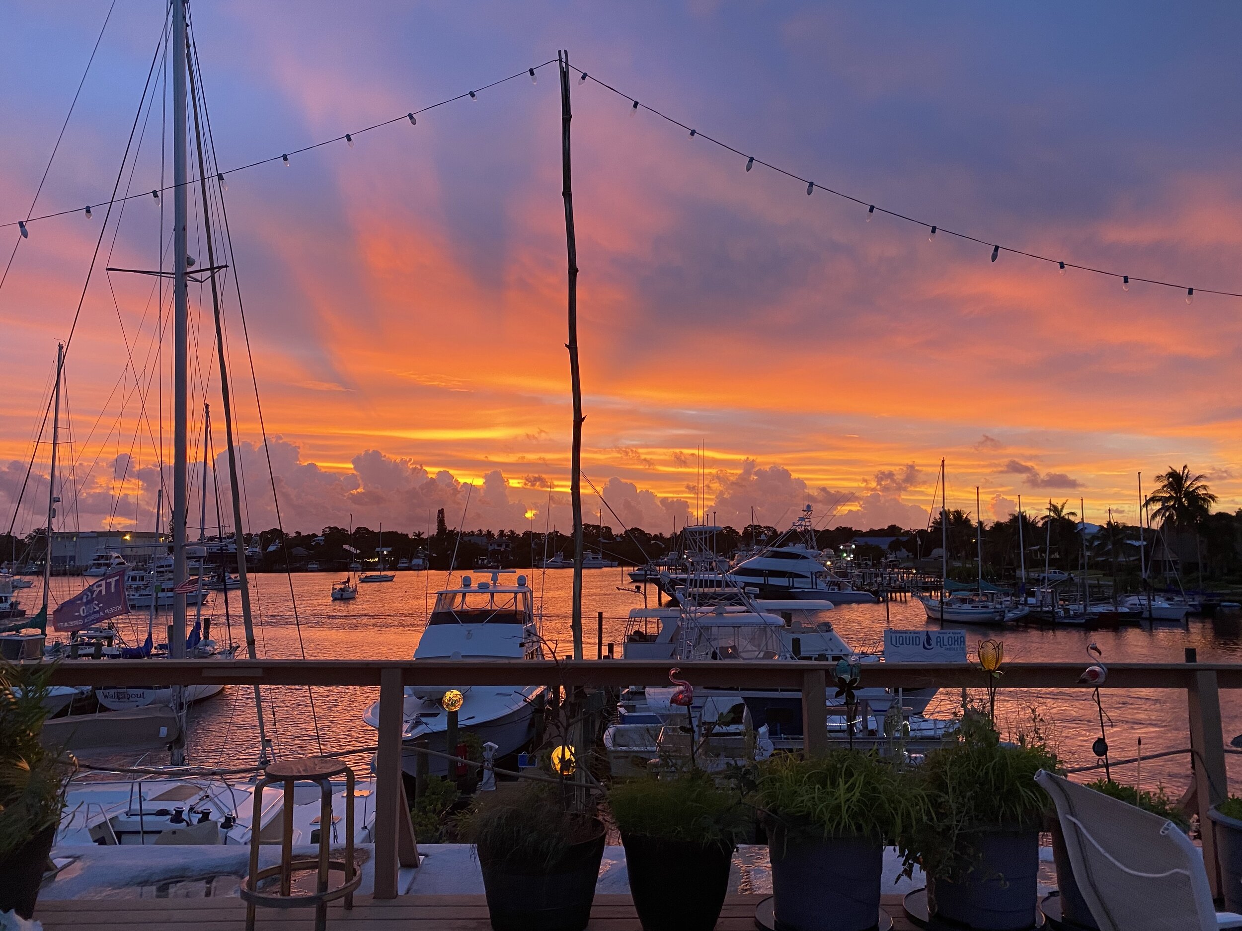 Sunset over a marina with sailboats and motor yachts moored in the harbor, seen from a dockside deck.