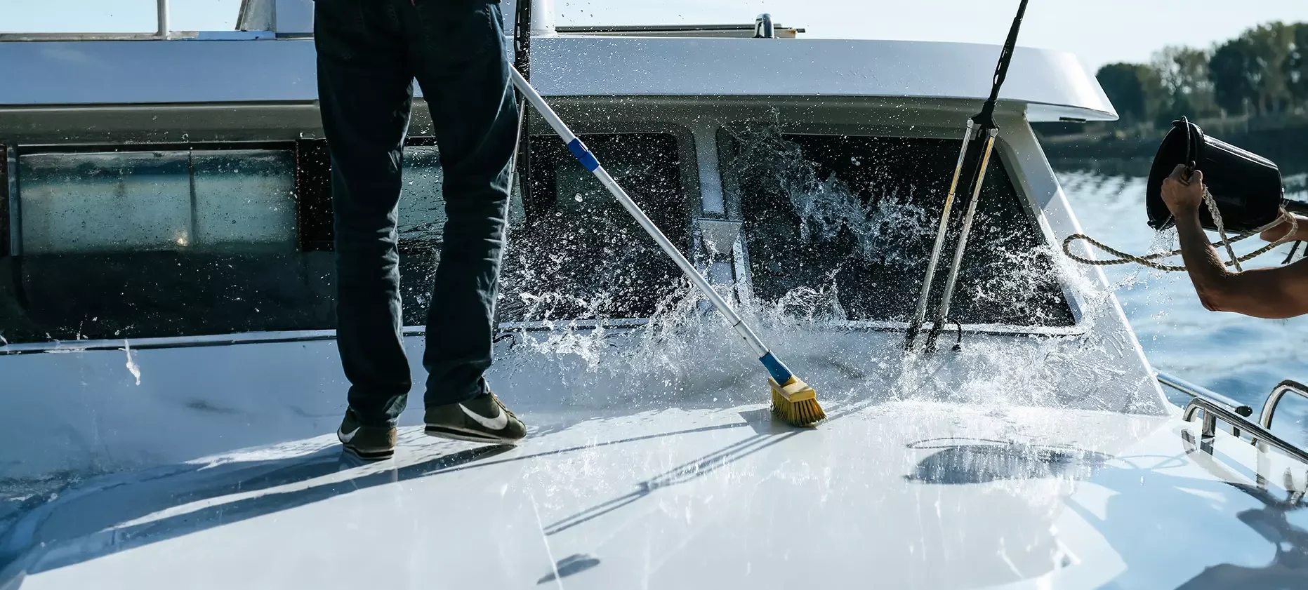 A crew member scrubbing a white motor yacht's deck and cabin windows with a long-handled brush and a bucket of water on the water.