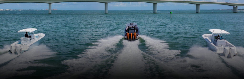 Three white center-console boats running in formation with wake trails beneath a bridge on open water.