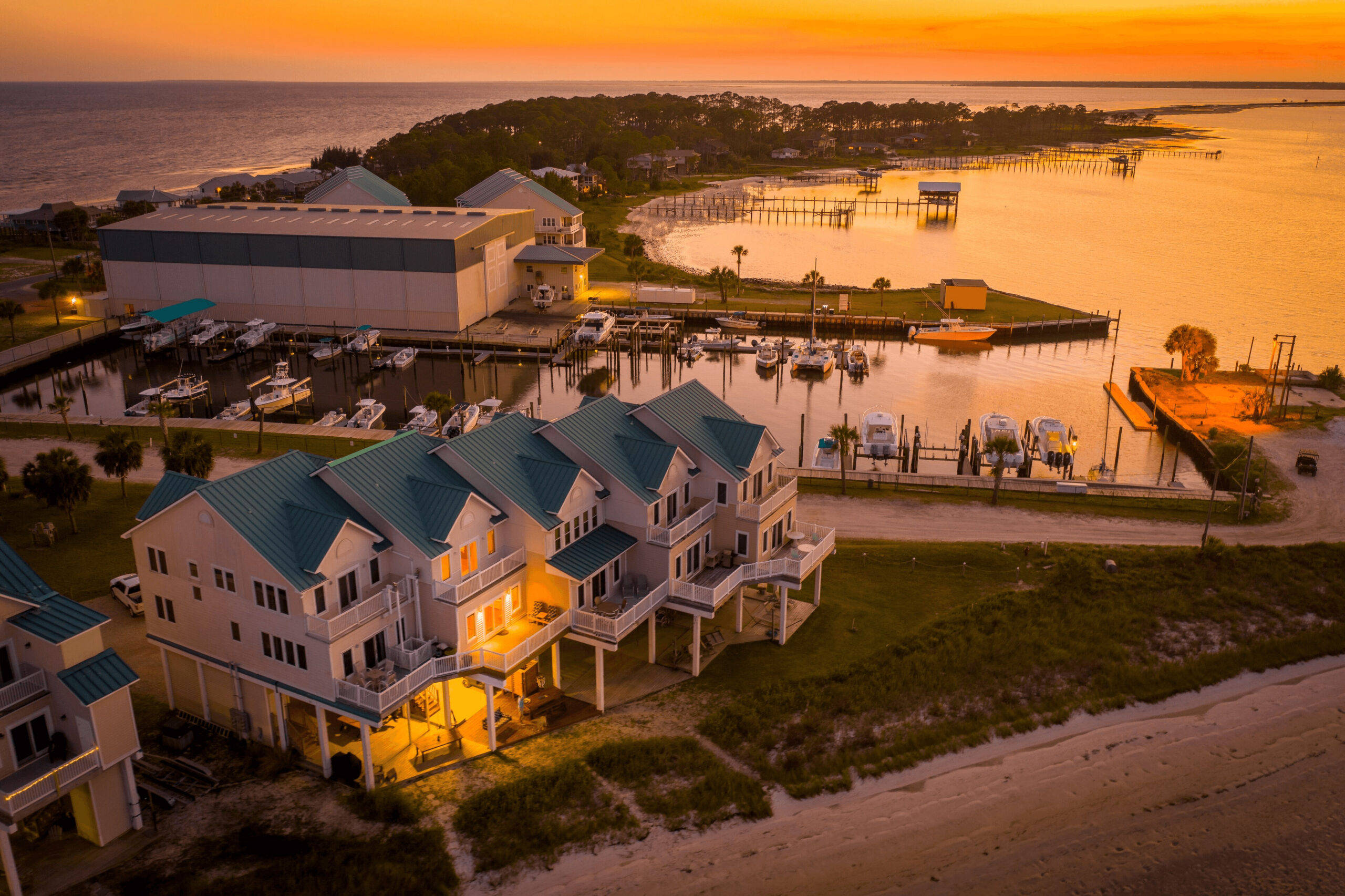 Aerial sunset view of boats moored at a waterfront marina beside a large boat storage building and beachfront homes, town unknown.