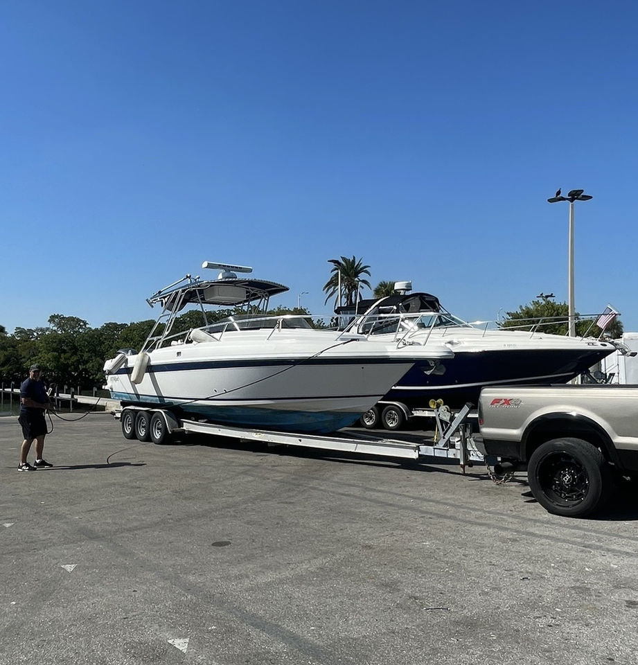 White and navy cabin cruiser on a trailer being rinsed by a man with a hose in a parking lot, with a pickup truck hitched behind.