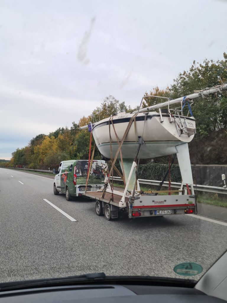 Sailboat secured on a flatbed trailer behind a green van on a motorway, with the trailer plate reading HEI C 143.