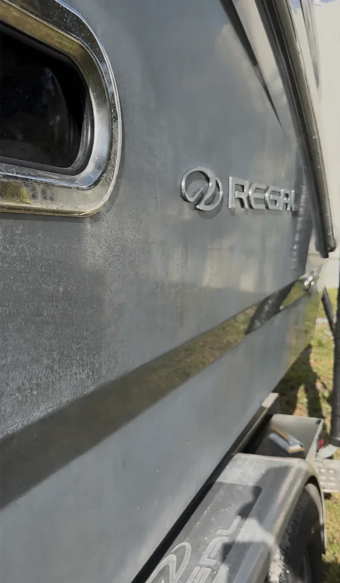 Side of a Regal boat hull with its chrome badge and porthole window, resting on a trailer outdoors.