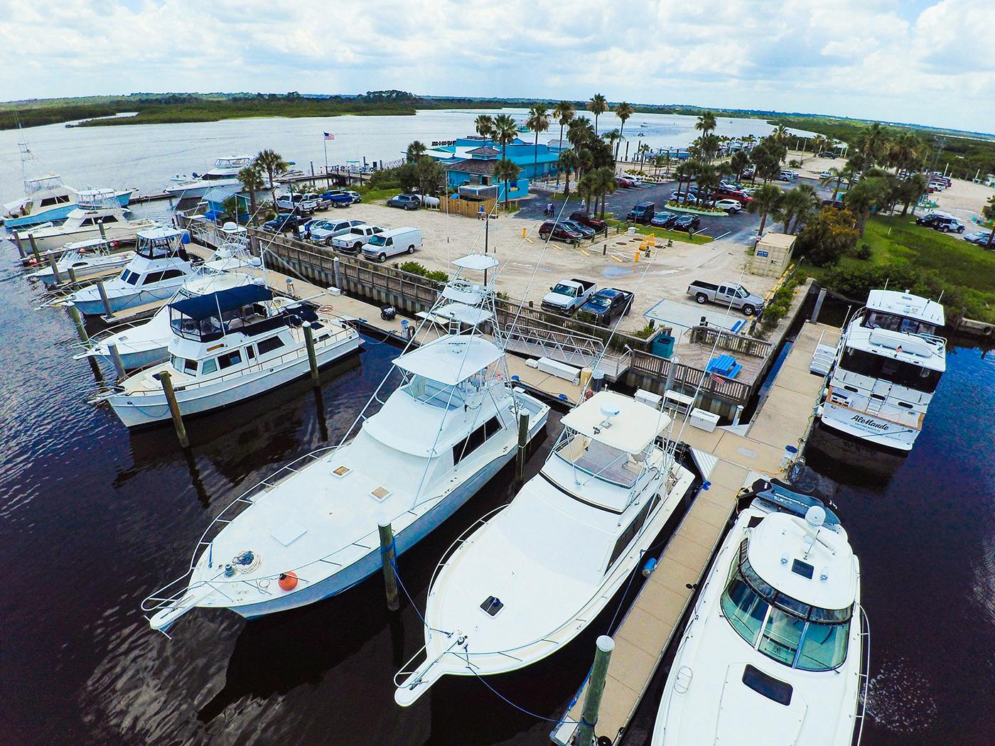 Aerial view of sport fishing boats docked at a marina with a waterfront parking lot and palm-lined shoreline, business and town not identified.