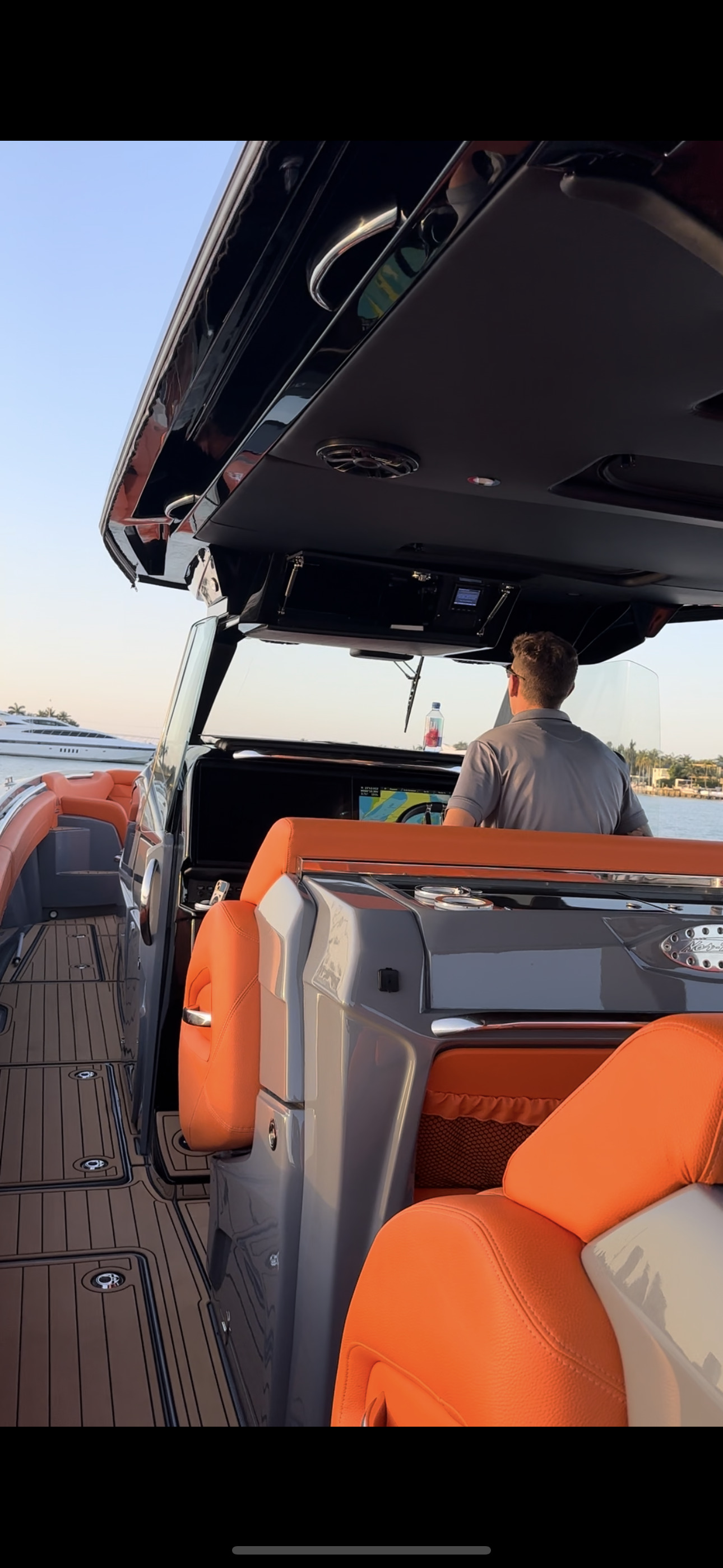 Luxury speedboat cockpit with orange leather seats and a helmsman at the console, underway near a moored white yacht and waterfront homes.
