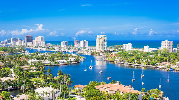 Aerial view of yachts and sailboats moored along a residential canal with high-rise condos on the skyline, likely Fort Lauderdale, Florida.