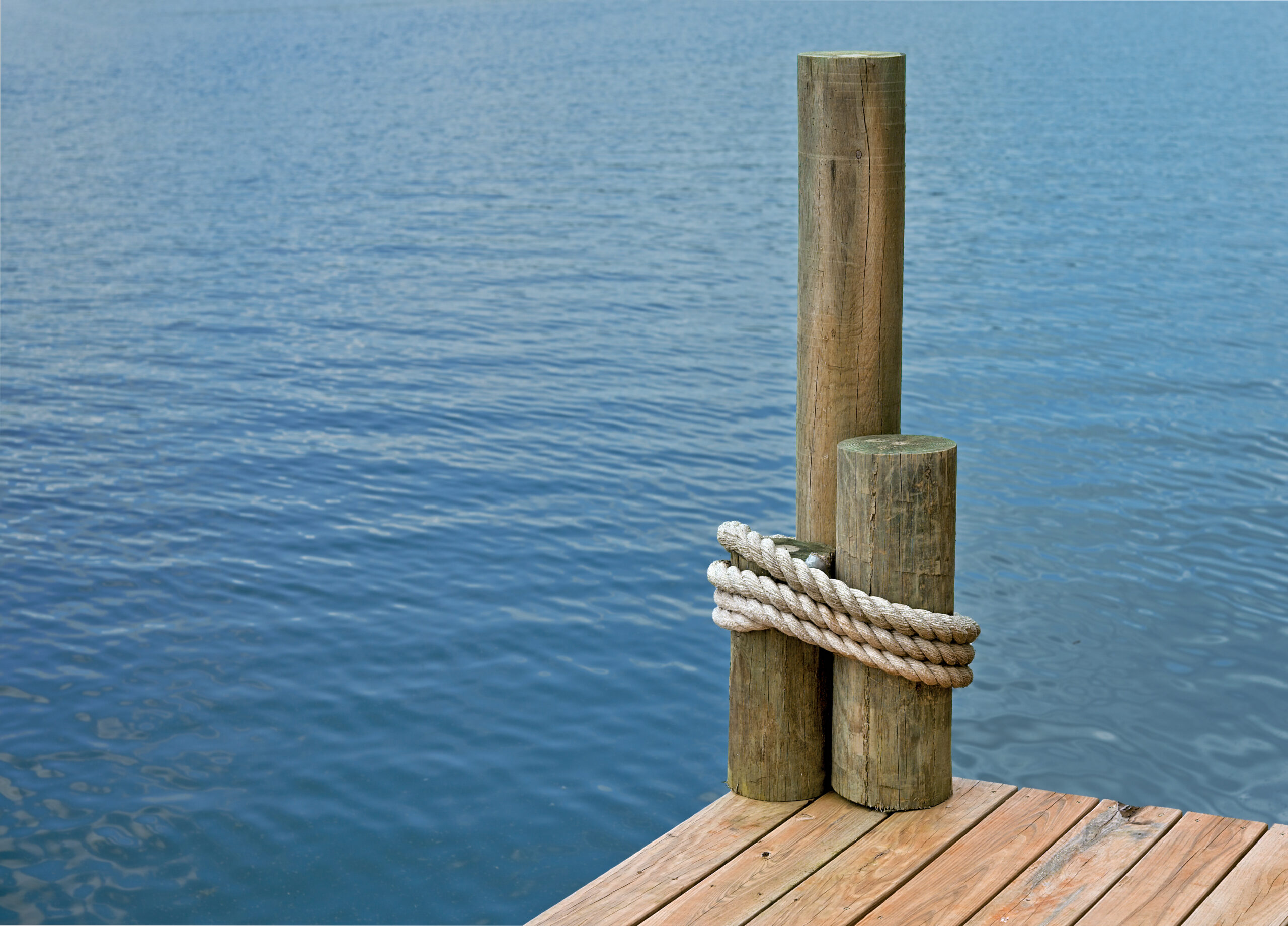 Wooden dock pilings with a thick rope tied around them at the edge of a calm lake or harbor.