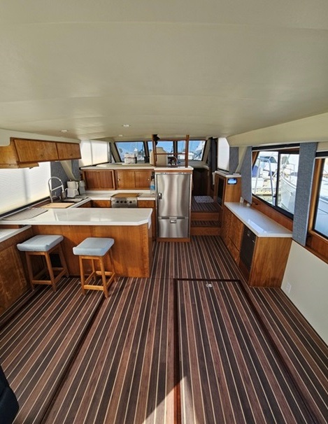 Interior salon of a motor yacht showing teak galley cabinetry, a breakfast bar with stools, and striped wood-look deck flooring.