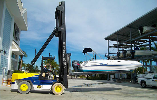 Yellow and blue forklift with a telehandler-style boom lifting a boat toward a covered dry-storage rack at a marina, with a white pickup parked nearby.