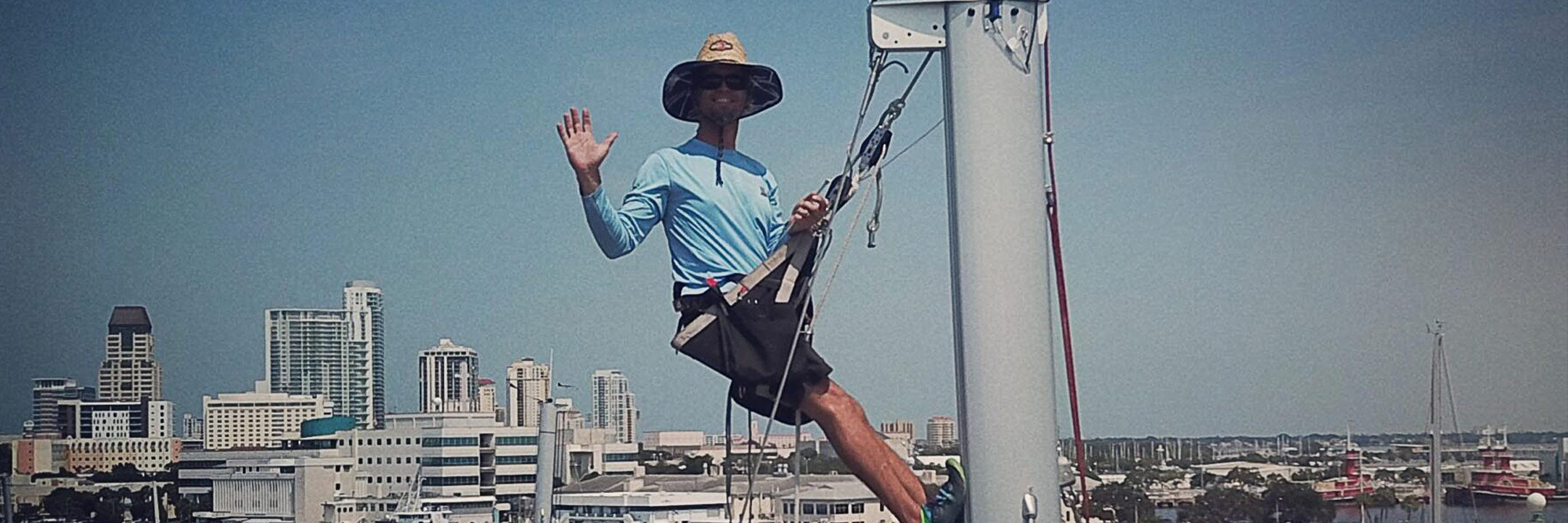 A rigger in a bosun's chair waves from a sailboat mast, with a downtown waterfront skyline behind him.