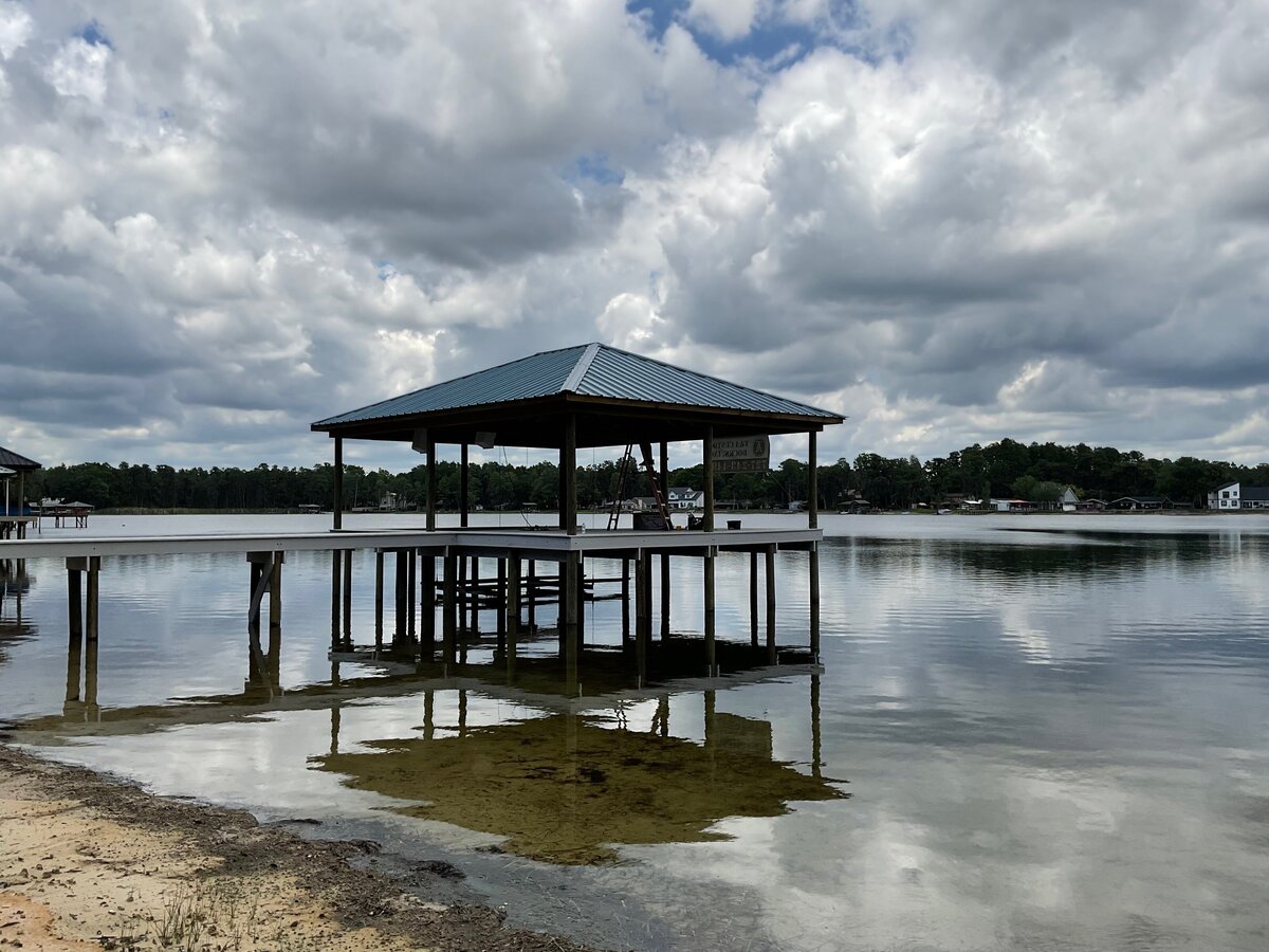 Covered wooden boat dock with a metal roof extending over calm lake water, with a sandy shore and lakeside homes in the distance.