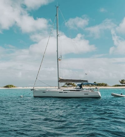 White sailboat at anchor in turquoise water beside a sandy island with palm trees under a partly cloudy sky.