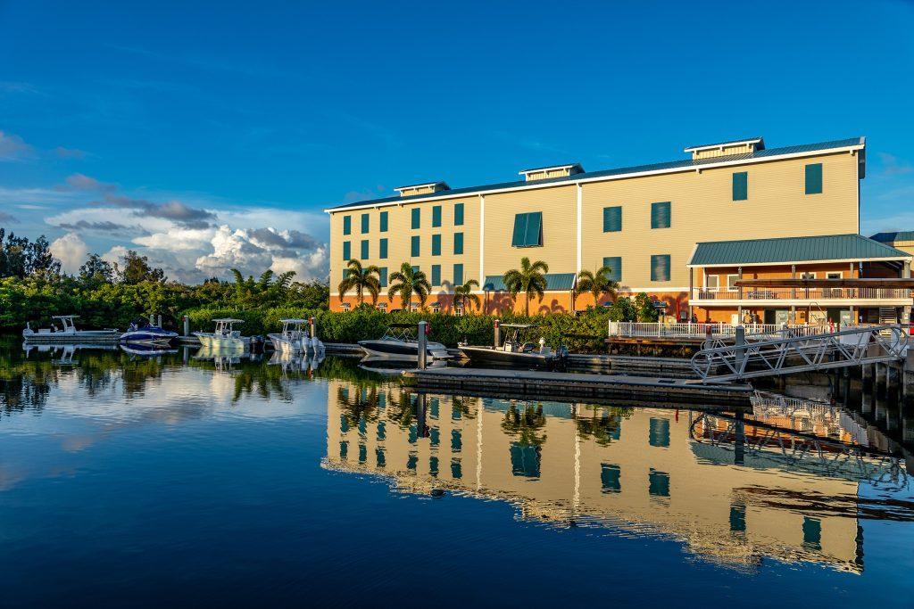 Waterfront hotel building with moored powerboats and a floating dock along a calm canal, with palm trees and blue sky.