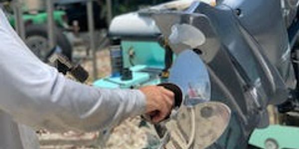 A person's hand grips the tiller of a grey outboard motor, with a teal engine stand and boat equipment in the background.