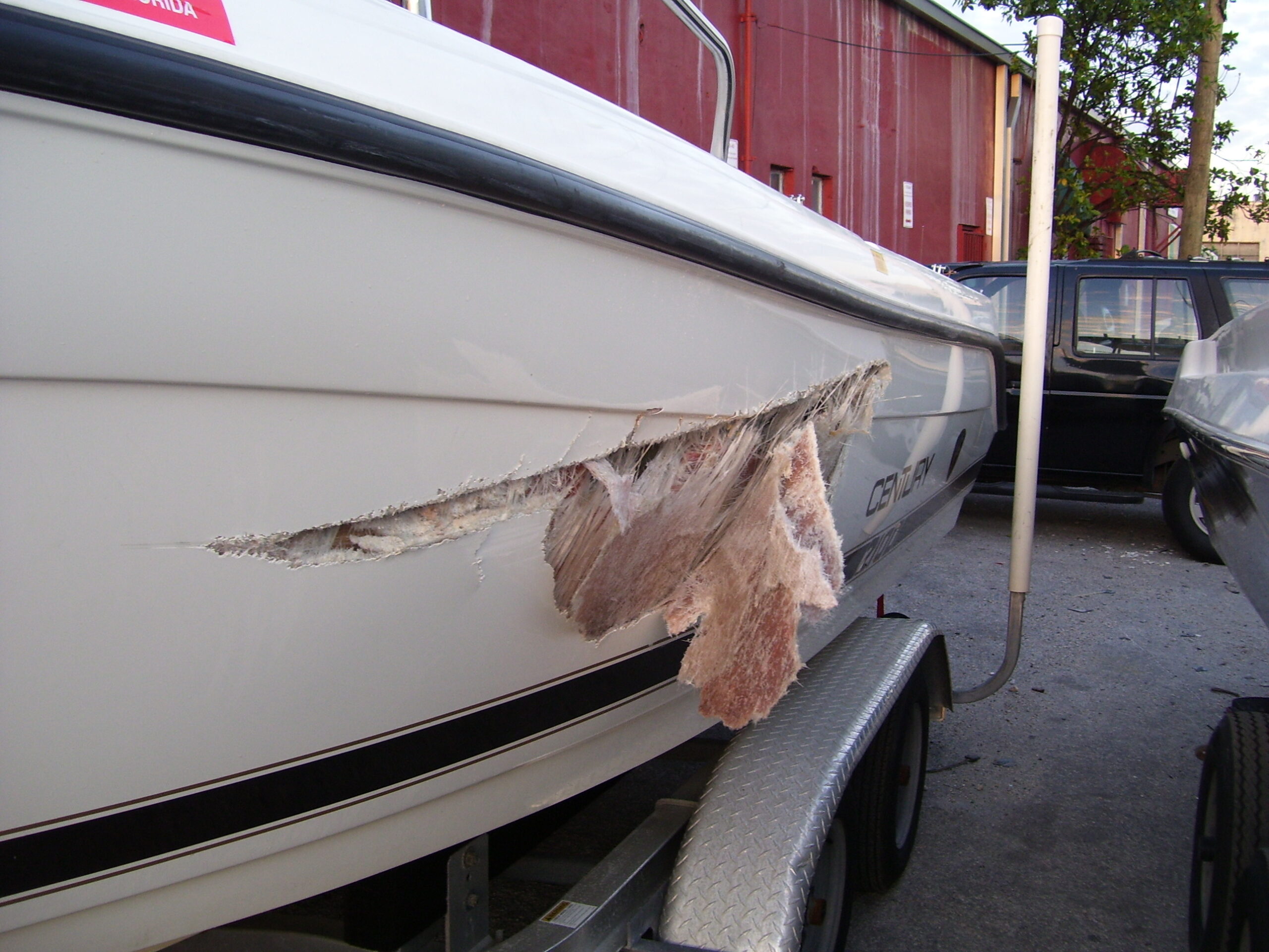 A white boat with a torn-open fiberglass hull and exposed laminate sitting on a trailer in a parking area, with a red building and a Florida sign in the background.