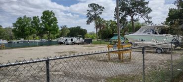 Gravel storage lot with RVs and a boat on a trailer behind a chain-link fence, with trees in the background.