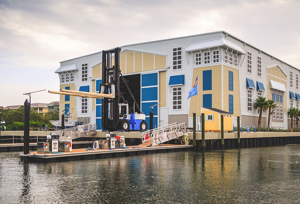 Large forklift handling a timber beam at the open bay doors of a waterfront boat storage building with a floating dock and boats along the canal.