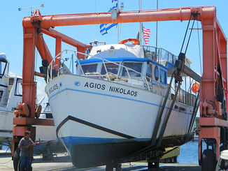 Blue-and-white boat Agios Nikolaos being hauled out by a travel lift at a boatyard.